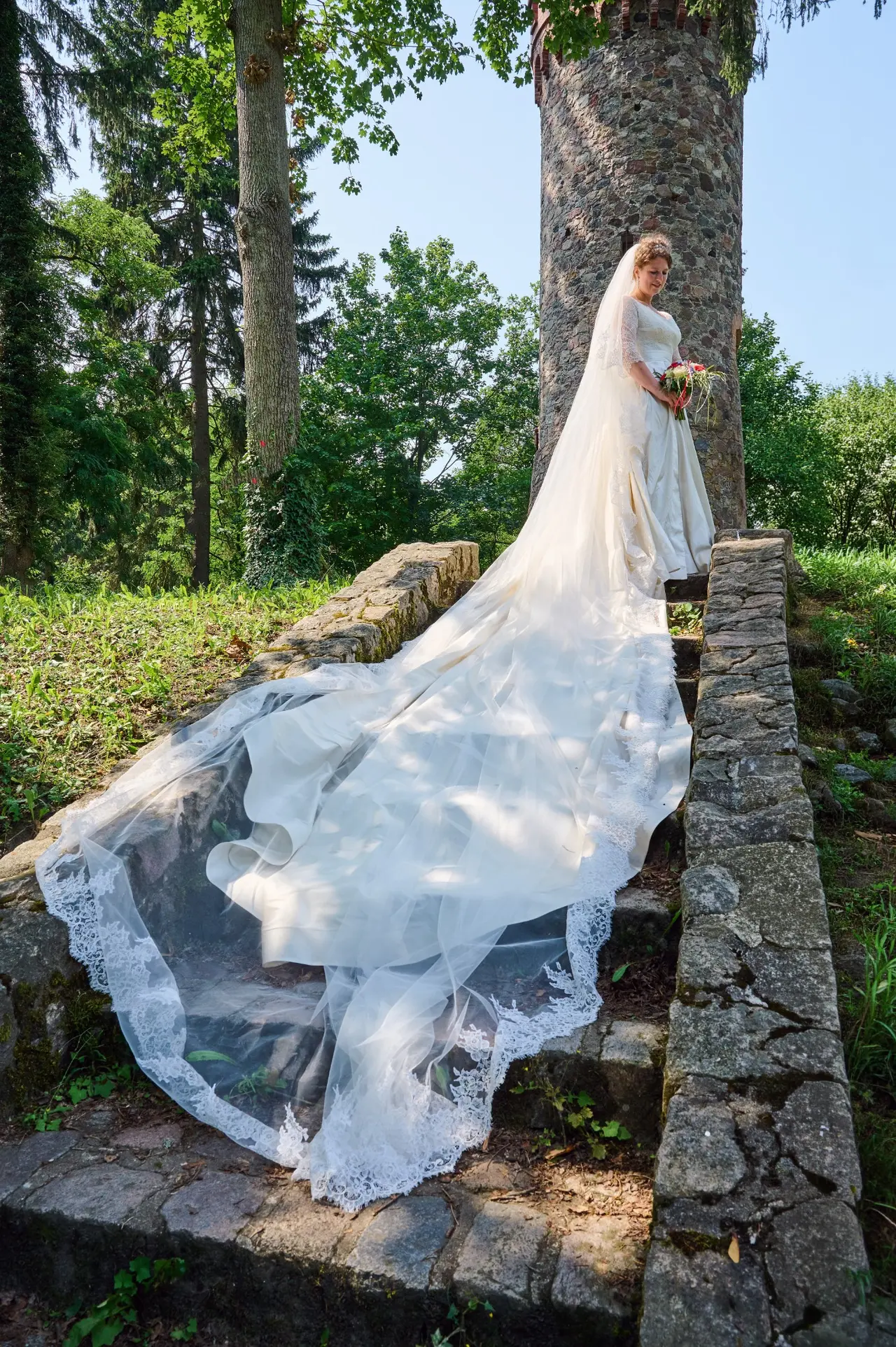 Braut im seidenen FRILISA Brautkleid mit langer Schleppe und Spitzenschleier auf einer Steintreppe vor einem Turm